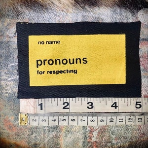 May include: Black fabric patch with a yellow rectangle that says "no name pronouns for respecting" in black text.