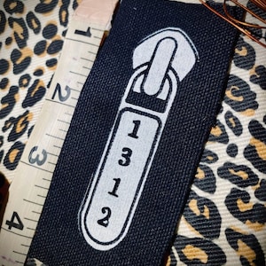 Könnte beinhalten: Ein schwarzes Stoffpatch mit einem weißen Reißverschluss-Design und den Zahlen "1312". Ein beiges Maßband mit schwarzen Markierungen ist teilweise sichtbar. Der Hintergrund zeigt ein Leopardenmuster.