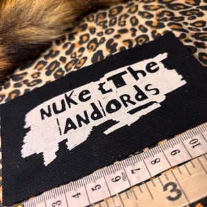 Peut inclure: &Eacute;cusson rectangulaire noir avec le texte "Nuke the Landlords" en blanc. Un m&egrave;tre ruban est plac&eacute; &agrave; c&ocirc;t&eacute; de l'&eacute;cusson. Le fond est un motif l&eacute;opard.