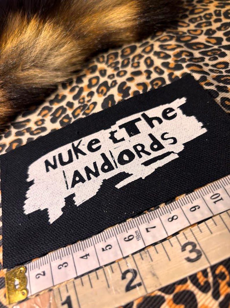 Peut inclure: &Eacute;cusson rectangulaire noir avec le texte "Nuke the Landlords" en blanc. Un m&egrave;tre ruban est plac&eacute; &agrave; c&ocirc;t&eacute; de l'&eacute;cusson. Le fond est un motif l&eacute;opard.
