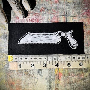 Könnte beinhalten: Schwarzer Stoff-Patch mit einer weißen Umrandung einer Säge mit einem Holzgriff.