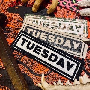 May include: Three fabric patches with the word "TUESDAY" printed in black and white on a beige background. The patches are stacked on top of each other, with the largest patch at the bottom. The patches are surrounded by a colourful fabric with a paisley pattern.