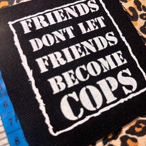 May include: A square black fabric patch with white screen-printed text that reads "FRIENDS DONT LET FRIENDS BECOME COPS" inside a white border. A blue measuring tape is visible on the left side, showing measurements in inches.