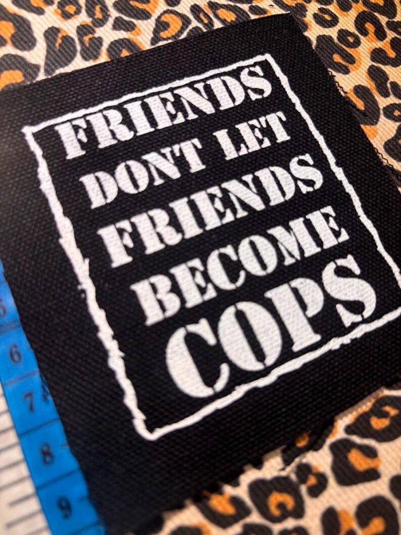 May include: A square black fabric patch with white screen-printed text that reads "FRIENDS DONT LET FRIENDS BECOME COPS" inside a white border. A blue measuring tape is visible on the left side, showing measurements in inches.