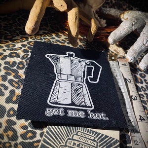 May include: Black fabric patch with a white illustration of a coffee pot and the text "get me hot."