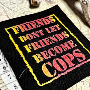 May include: A square black fabric patch featuring the yellow and red gradient text "FRIENDS DONT LET FRIENDS BECOME COPS" enclosed in a jagged yellow border. A white measuring tape is visible along the left edge, showing a length of 3.5 inches.