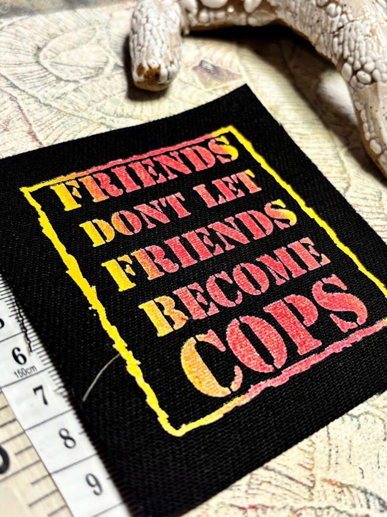 May include: A square black fabric patch featuring the yellow and red gradient text "FRIENDS DONT LET FRIENDS BECOME COPS" enclosed in a jagged yellow border. A white measuring tape is visible along the left edge, showing a length of 3.5 inches.