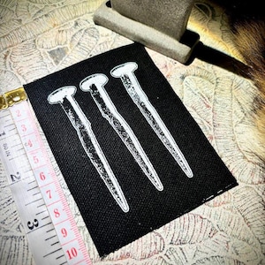 以下が含まれることがあります： 3つの釘の白いイラストが描かれた黒いパッチ。メジャーテープでパッチの長さが約10cmであることが示されています。パッチは模様のある表面にあります。