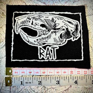 Pode incluir: Patch bordado em preto e branco com uma caveira de rato e a palavra "RAT" em letras brancas. O patch mede aproximadamente 5 cm de largura.