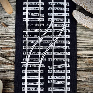 May include: A black fabric patch with a white graphic of a railroad track switch. The track is made up of white lines that represent the wooden ties and rails.