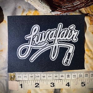 Puede incluir: Un parche de tela negro con bordado blanco. El parche presenta la palabra "Lavafair" en una fuente de letra cursiva estilizada con un contorno blanco. Un contorno blanco de un conector de cable de micrófono se encuentra debajo de la palabra.