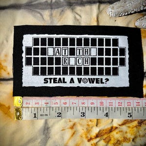 Op de afbeelding: Een zwart-wit patch met de tekst "AT TH RCH STEAL A VOWEL?" in een kruiswoordpuzzelstijl. De patch bevindt zich op een tie-dye stof. Een meetlint geeft aan dat de patch ongeveer 13 cm lang is.