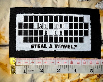 May include: A black and white patch with the text "AT TH RCH STEAL A VOWEL?" in a crossword puzzle style. The patch is on a tie-dye fabric. A measuring tape shows the patch is approximately 5 inches long.
