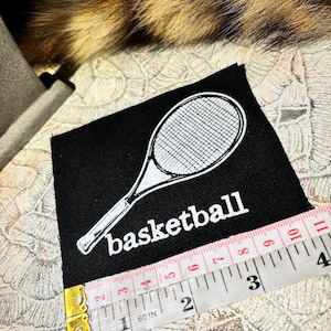 Puede incluir: Parche cuadrado negro con un diseño de raqueta de tenis blanca y la palabra "basketball" impresa debajo. Una cinta métrica muestra que el parche mide aproximadamente 10 cm de ancho.