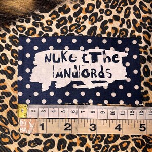 Peut inclure: Un &eacute;cusson bleu marine &agrave; pois blancs avec les mots "Nuke the Landlords" en blanc vieilli. L'&eacute;cusson mesure environ 5 cm de large. Le fond est un tissu &agrave; imprim&eacute; l&eacute;opard.