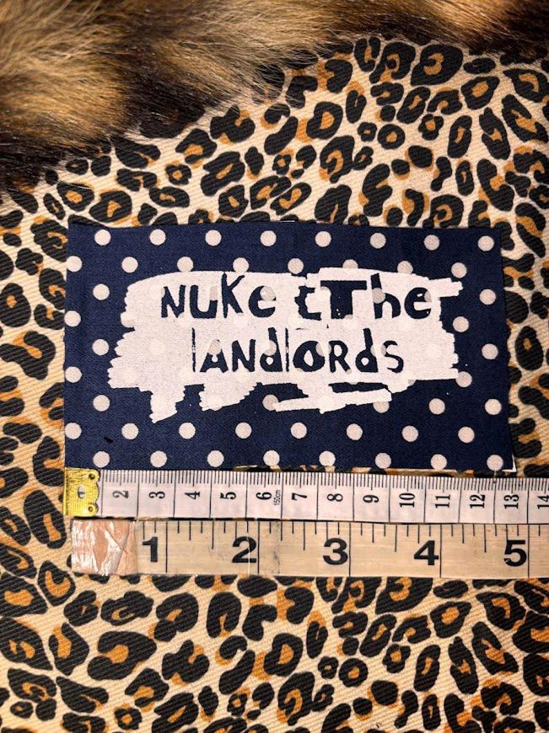 Peut inclure: Un &eacute;cusson bleu marine &agrave; pois blancs avec les mots "Nuke the Landlords" en blanc vieilli. L'&eacute;cusson mesure environ 5 cm de large. Le fond est un tissu &agrave; imprim&eacute; l&eacute;opard.