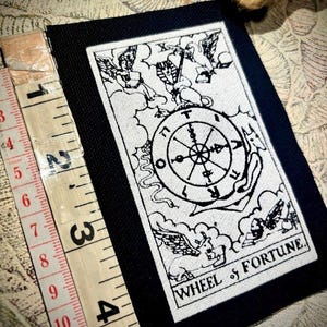 Puede incluir: Un parche de carta del tarot en blanco y negro con la Rueda de la Fortuna. El parche tiene un borde negro y el texto "WHEEL of FORTUNE". Una cinta métrica indica que el parche mide unos 10 cm de largo.