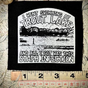 May include: Black square patch with white text and a graphic of a lake and mountains. The text reads "I WENT SWIMMING AT TROUT LAKE AND ALL I GOT WAS THIS STAPH INFECTION". The patch is approximately 4 inches wide.