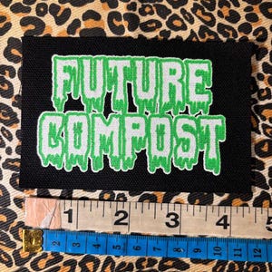 May include: A rectangular black embroidered patch featuring the text FUTURE COMPOST in a dripping font with silver lettering and a bright green outline. A measuring tape is positioned below the patch, showing a width of approximately 4 inches.