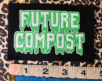 Puede incluir: Un parche rectangular negro bordado con el texto FUTURE COMPOST en una fuente plateada que gotea con un contorno verde brillante. Una cinta métrica colocada debajo del parche muestra un ancho de aproximadamente 11,5 cm.