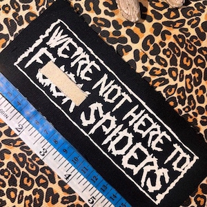 May include: A rectangular black fabric patch featuring white embroidered text that reads "WE'RE NOT HERE TO F*** SPIDERS". A white measuring tape is positioned along the edge, showing a length of approximately 15 cm for the patch.