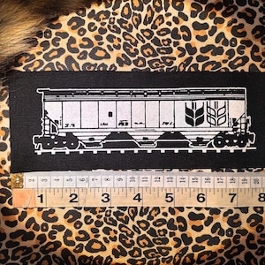 May include: Black rectangular patch featuring a white illustration of a train car with a wheat symbol. The patch is on a leopard print background. A ruler shows measurements in inches.