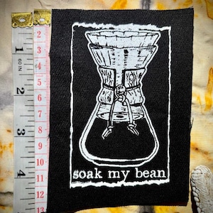 Può includere: Una toppa nera con una grafica bianca di una caffettiera e la scritta "soak my bean". La toppa misura circa 10 cm di altezza e 7,6 cm di larghezza. I bordi della toppa hanno un aspetto sfilacciato e usurato.