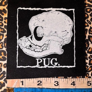 May include: A square black fabric patch featuring a white screen-printed illustration of a pug skull inside a rough-edged border with the text PUG. below it. A measuring tape shows the patch is approximately 4.5 inches wide at the base.