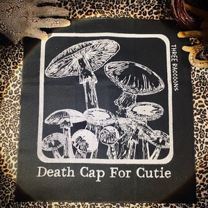 May include: A black fabric patch featuring a white screen-printed illustration of various mushrooms within a rounded rectangular border. The text "THREE RACCOONS" is printed vertically on the right, and "Death Cap For Cutie" is printed below.