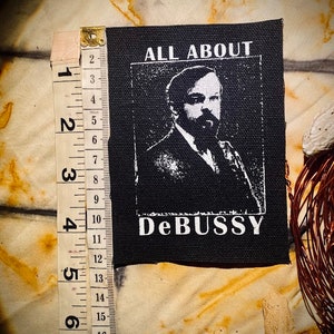 Peut inclure: Patch thermocollant noir et blanc avec un portrait d'un homme et le texte "ALL ABOUT DEBUSSY".