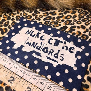 Peut inclure: Un tissu bleu marine &agrave; pois blancs avec le texte "NUKE THE LANDLORDS" en blanc. Une r&egrave;gle en bois est plac&eacute;e en diagonale sur le tissu. Le fond pr&eacute;sente un motif l&eacute;opard.