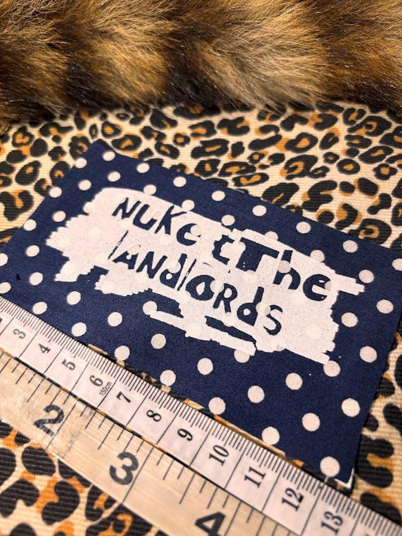 Peut inclure: Un tissu bleu marine &agrave; pois blancs avec le texte "NUKE THE LANDLORDS" en blanc. Une r&egrave;gle en bois est plac&eacute;e en diagonale sur le tissu. Le fond pr&eacute;sente un motif l&eacute;opard.