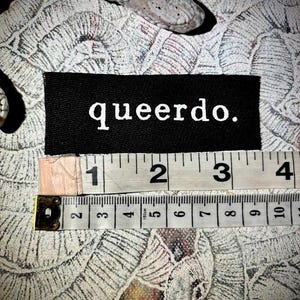 May include: Black fabric patch with white text that reads "queerdo." The patch is about 2 inches wide and 1 inch tall. A measuring tape is visible below the patch.