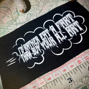 Pode incluir: Um patch retangular preto com texto branco que diz "I'D RATHER EAT A FART THAN BUY YOUR AI ART" em um design de nuvem. Uma fita métrica amarela e branca está ao lado do patch, mostrando centímetros.