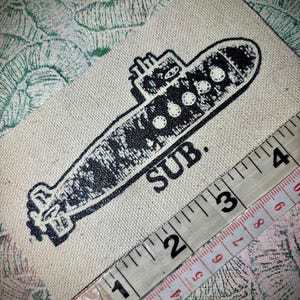 May include: A rectangular off-white fabric patch featuring a black screen-printed illustration of a submarine with the text SUB. positioned below it. A measuring tape is placed diagonally across the patch, showing a length of approximately 10 cm.