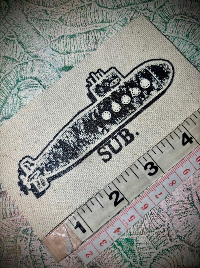 May include: A rectangular off-white fabric patch featuring a black screen-printed illustration of a submarine with the text SUB. positioned below it. A measuring tape is placed diagonally across the patch, showing a length of approximately 10 cm.