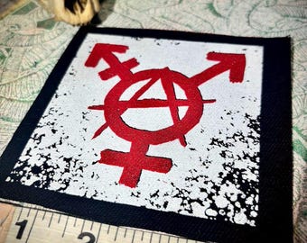 May include: Square patch with a black border and a white center featuring a red transgender anarchy symbol. The patch is approximately 4 inches wide. A measuring tape is placed below the patch.