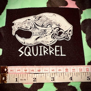 Op de afbeelding: Zwarte stoffen patch met een witte omtrek van een eekhoornschedel en het woord "SQUIRREL" in witte tekst.