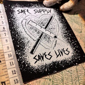 May include: Black and white patch with the text "SAFE SUPPLY SAVES LIVES". The patch features a heart shape with two crossed syringes. A measuring tape is visible on the left side.