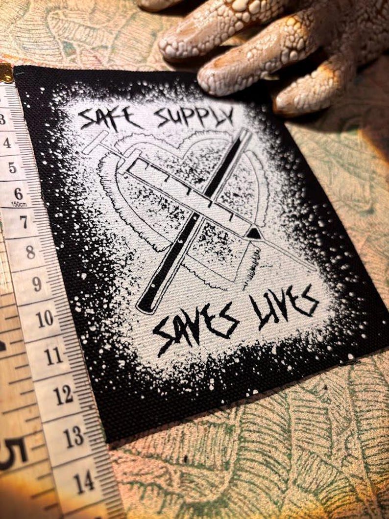May include: Black and white patch with the text "SAFE SUPPLY SAVES LIVES". The patch features a heart shape with two crossed syringes. A measuring tape is visible on the left side.