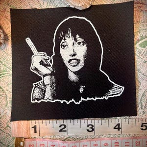 Op de afbeelding: Zwarte patch met een witte afbeelding van een vrouw die een mes vasthoudt, met een geschokte uitdrukking. De patch is ongeveer 10 cm breed. Een meetlint is eronder geplaatst.