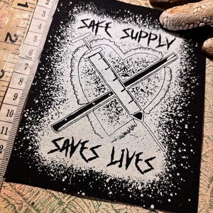 May include: Black and white patch with the text "SAFE SUPPLY SAVES LIVES". The design features a heart shape with two crossed syringes. The patch is approximately 10 cm wide.
