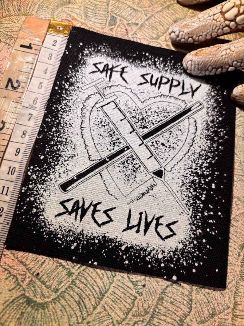 May include: Black and white patch with the text "SAFE SUPPLY SAVES LIVES". The design features a heart shape with two crossed syringes. The patch is approximately 10 cm wide.
