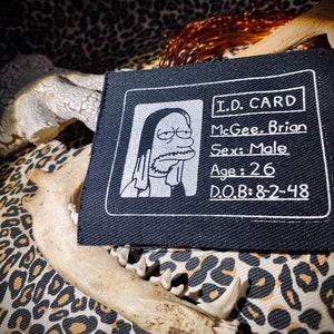 以下が含まれることがあります： ひげと長い髪を持つ人物の漫画の画像が描かれた白黒のIDカードパッチ。カードのテキストには、「I.D. CARD」、「McGee. Brian」、「Sex: Male」、「Age: 26」、「D.O.B: 8-2-48」と記載されています。