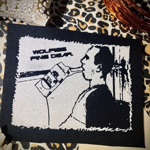 Può includere: Toppa ricamata in bianco e nero con una persona che beve da una bottiglia. Il testo "WOLFPACK FINE BEER" è visibile sulla toppa.