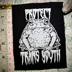 Può includere: Toppa quadrata nera con testo bianco e illustrazione di una rana. Il testo recita "PROTECT TRANS YOUTH". La toppa è alta circa 10 cm.