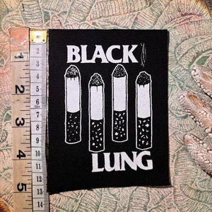 Könnte beinhalten: Schwarzer quadratischer Aufnäher mit den Worten "BLACK LUNG" in weißen Blockbuchstaben. Vier Illustrationen von weißen und schwarzen Zigaretten sind auf dem Aufnäher zentriert. Ein Maßband befindet sich auf der linken Seite des Aufnähers.