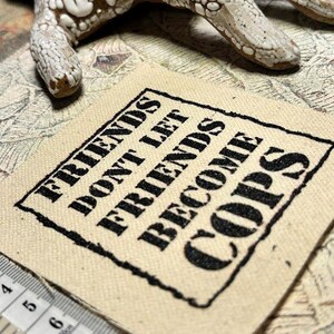 May include: A square beige fabric patch featuring the black embroidered text "FRIENDS DON'T LET FRIENDS BECOME COPS" enclosed in a rectangular border. A white measuring tape is visible at the bottom left, showing measurements in centimeters.