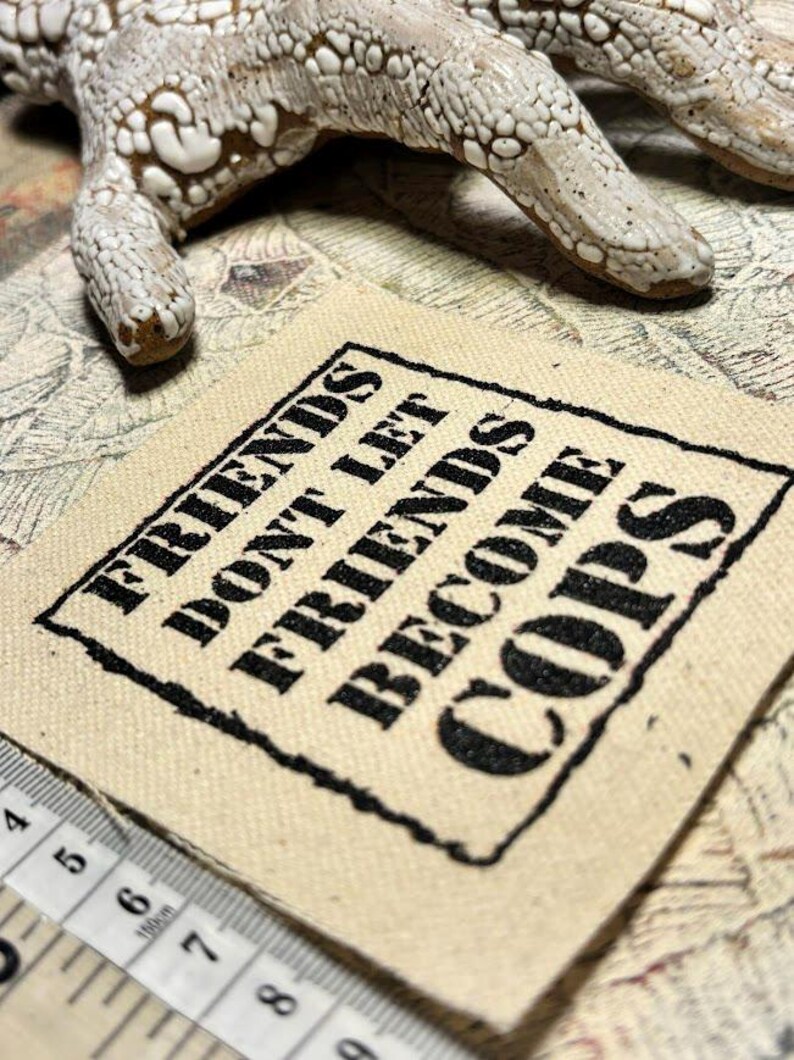 May include: A square beige fabric patch featuring the black embroidered text "FRIENDS DON'T LET FRIENDS BECOME COPS" enclosed in a rectangular border. A white measuring tape is visible at the bottom left, showing measurements in centimeters.