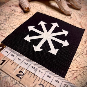 Op de afbeelding: Zwart vierkantige patch met een wit symbool van acht pijlen. Een meetlint geeft aan dat de patch ongeveer 7,6 cm breed is. De patch ligt op een patroon, met een decoratieve hand op de achtergrond.